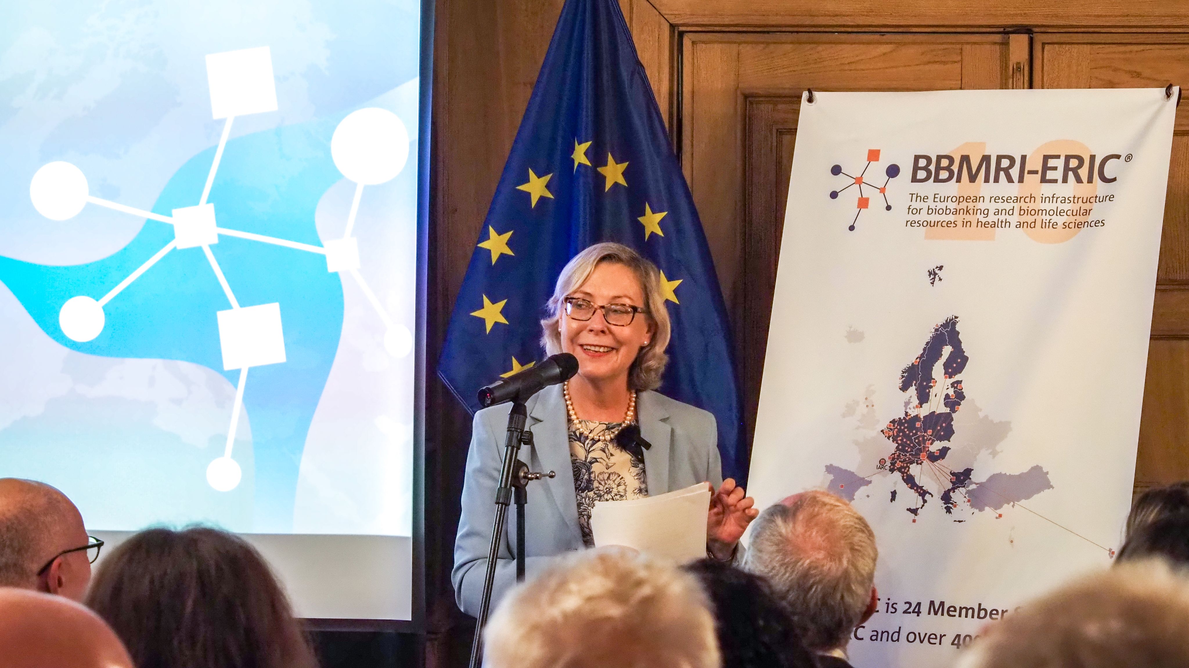 The picture of a white woman with blonde hair and glasses presenting to an audience. 