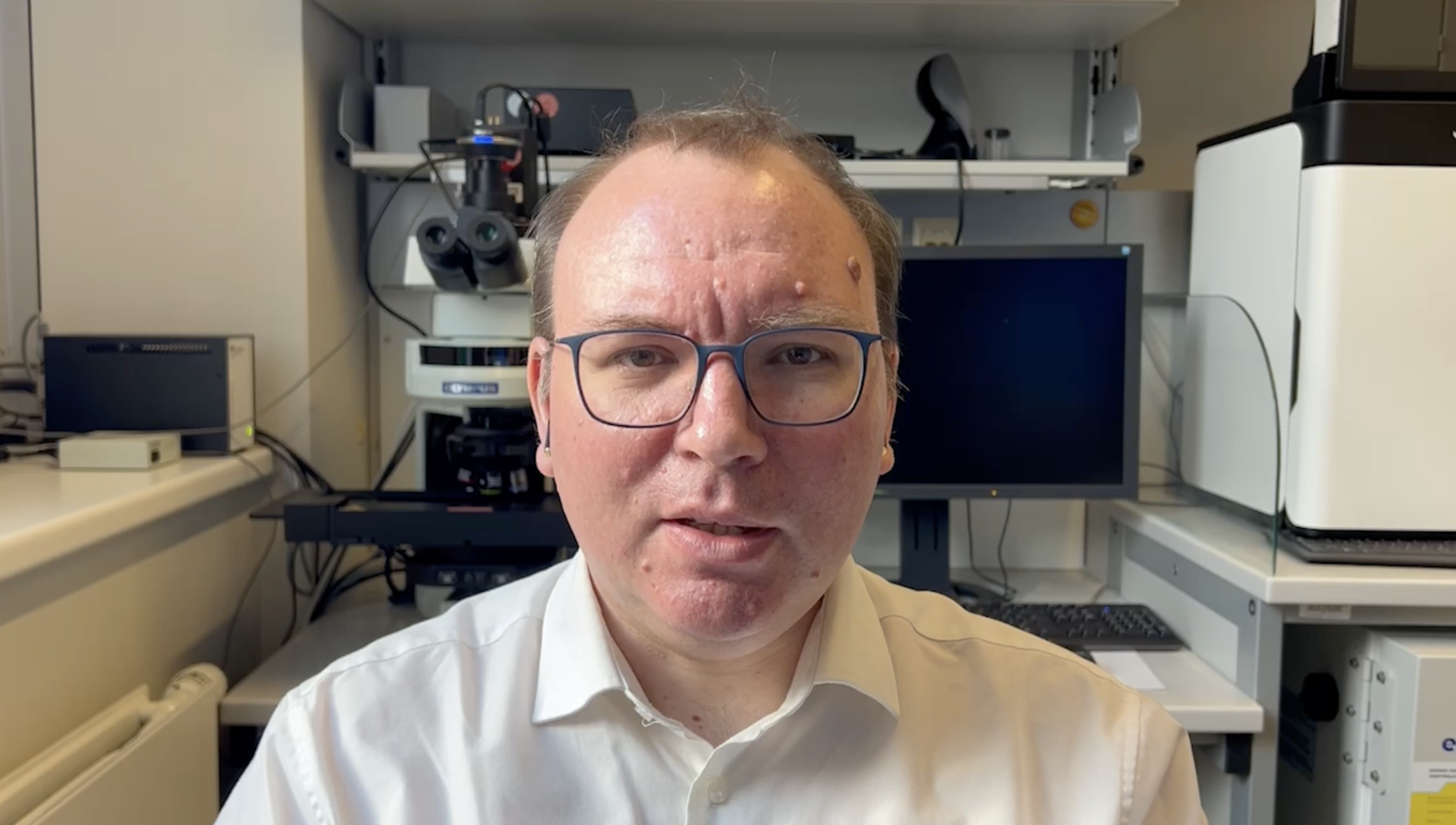 The picture of a white man wearing glasses. He is sitting in a laboratory and ihe is looking at the camera.