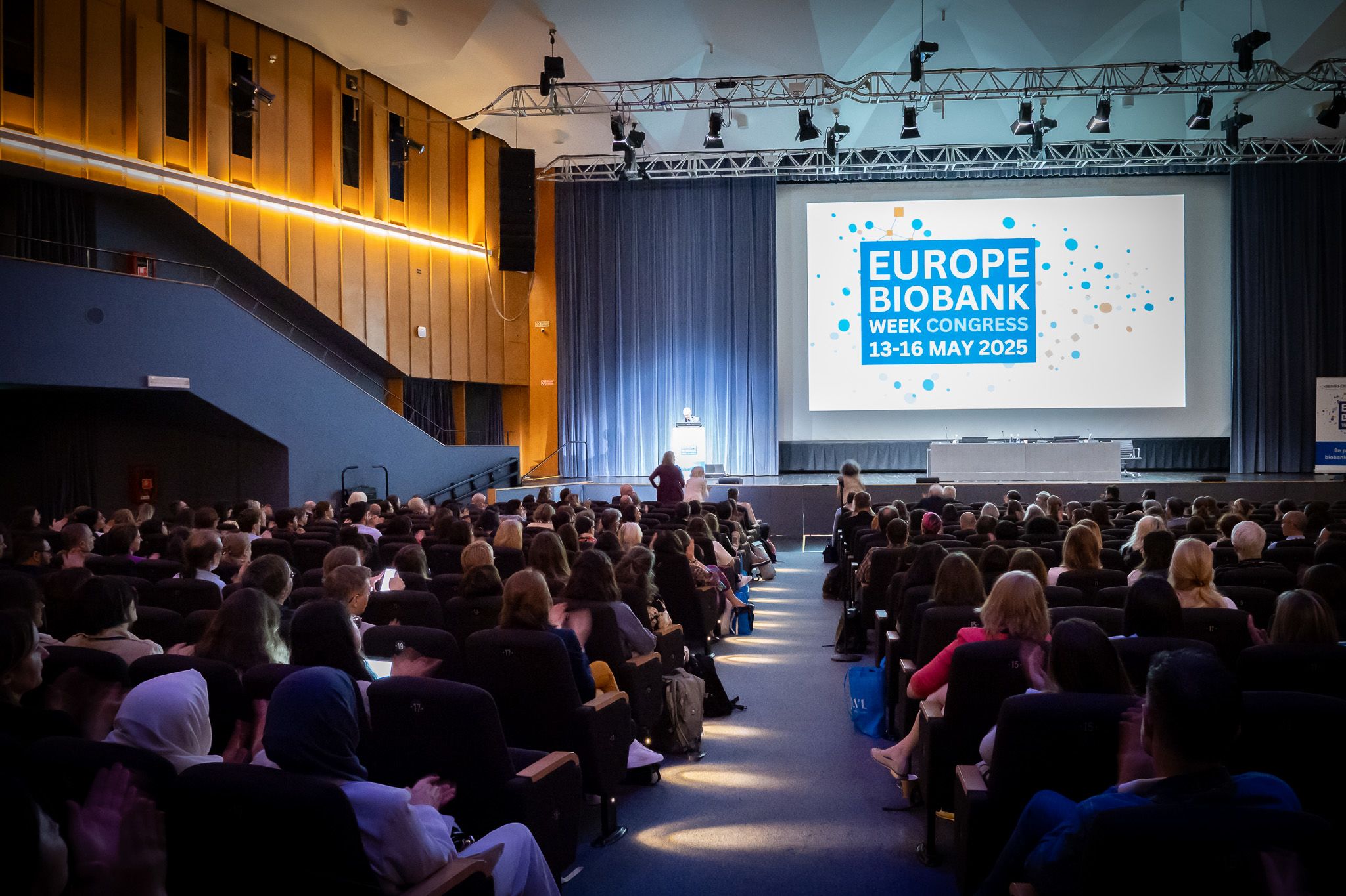 A view from the back of a large lecture hall showing a man at the podium and the large screen showing the EBW logo.
