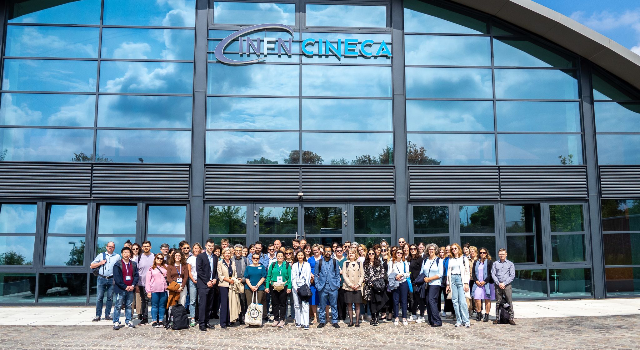 A large group of people posing in front of building with a large glass fassade that reflects the cloudy but bright sky.s