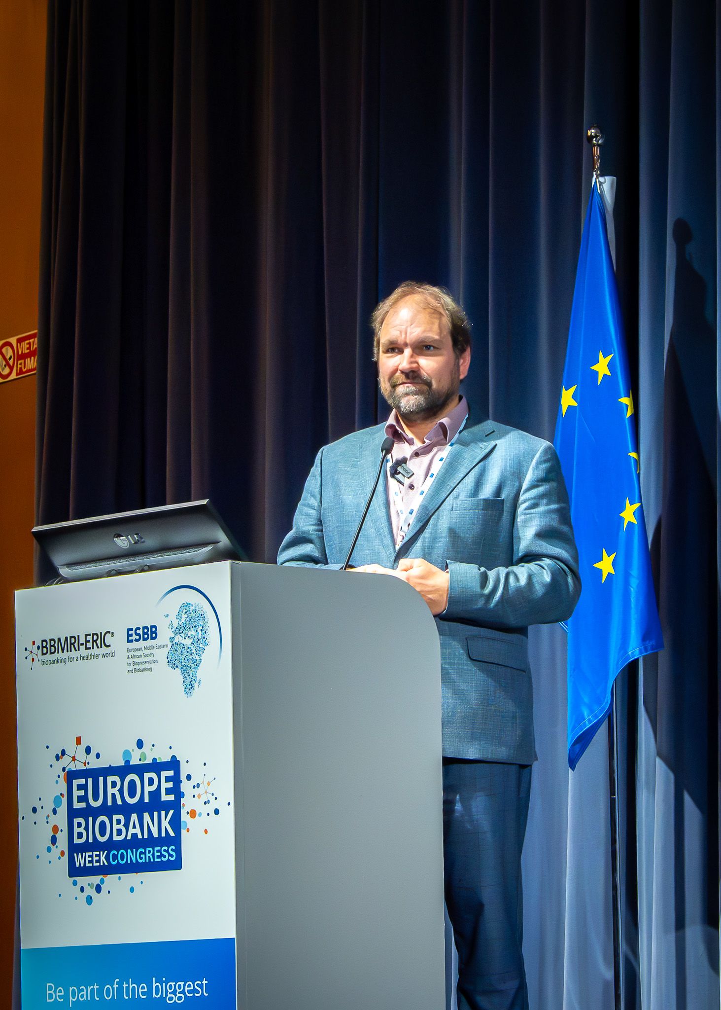 A close up of a man standing on a stage in front of a curtain and an EU flag.