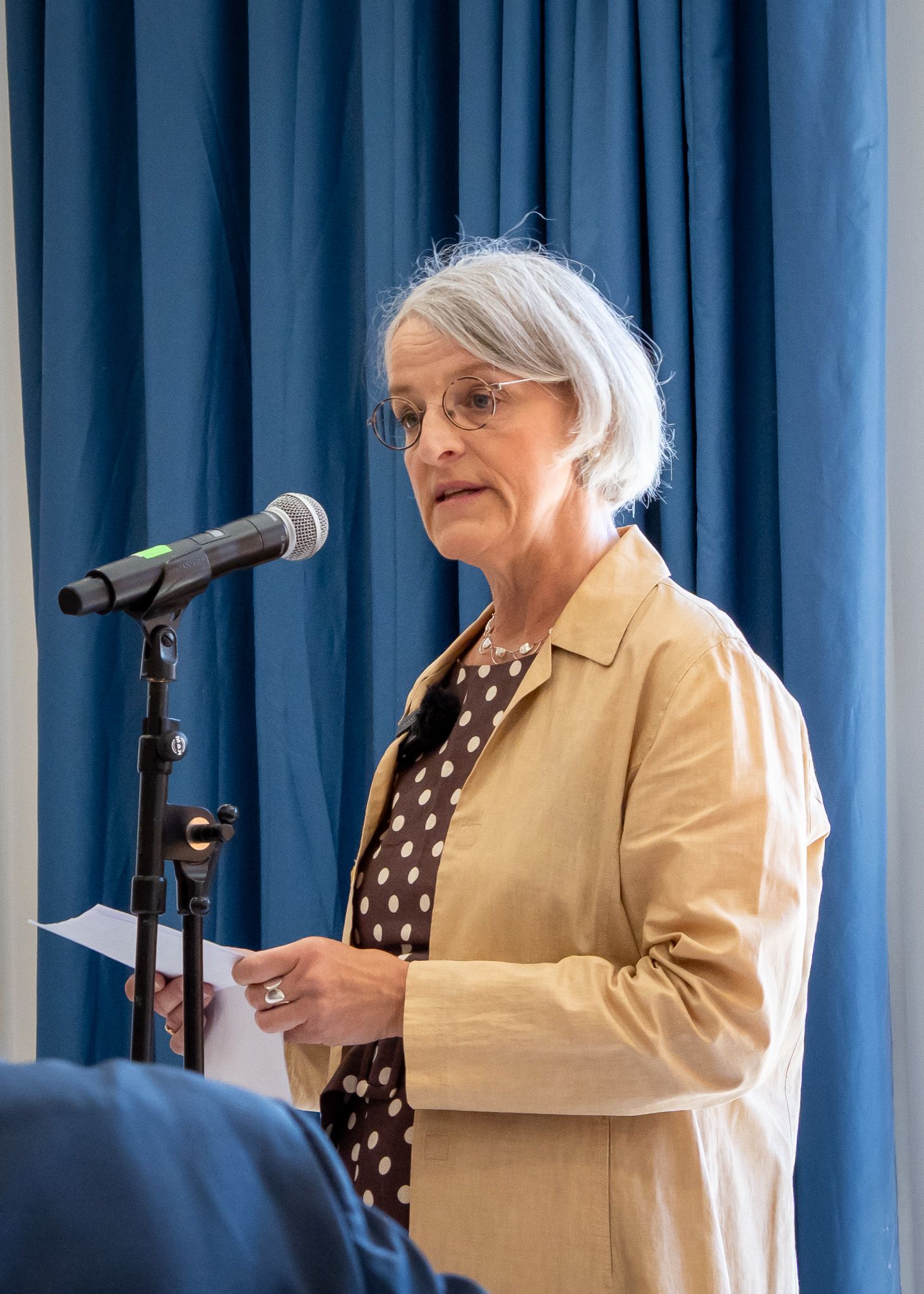 A woman in front of blue curtains speaking into a microphone.