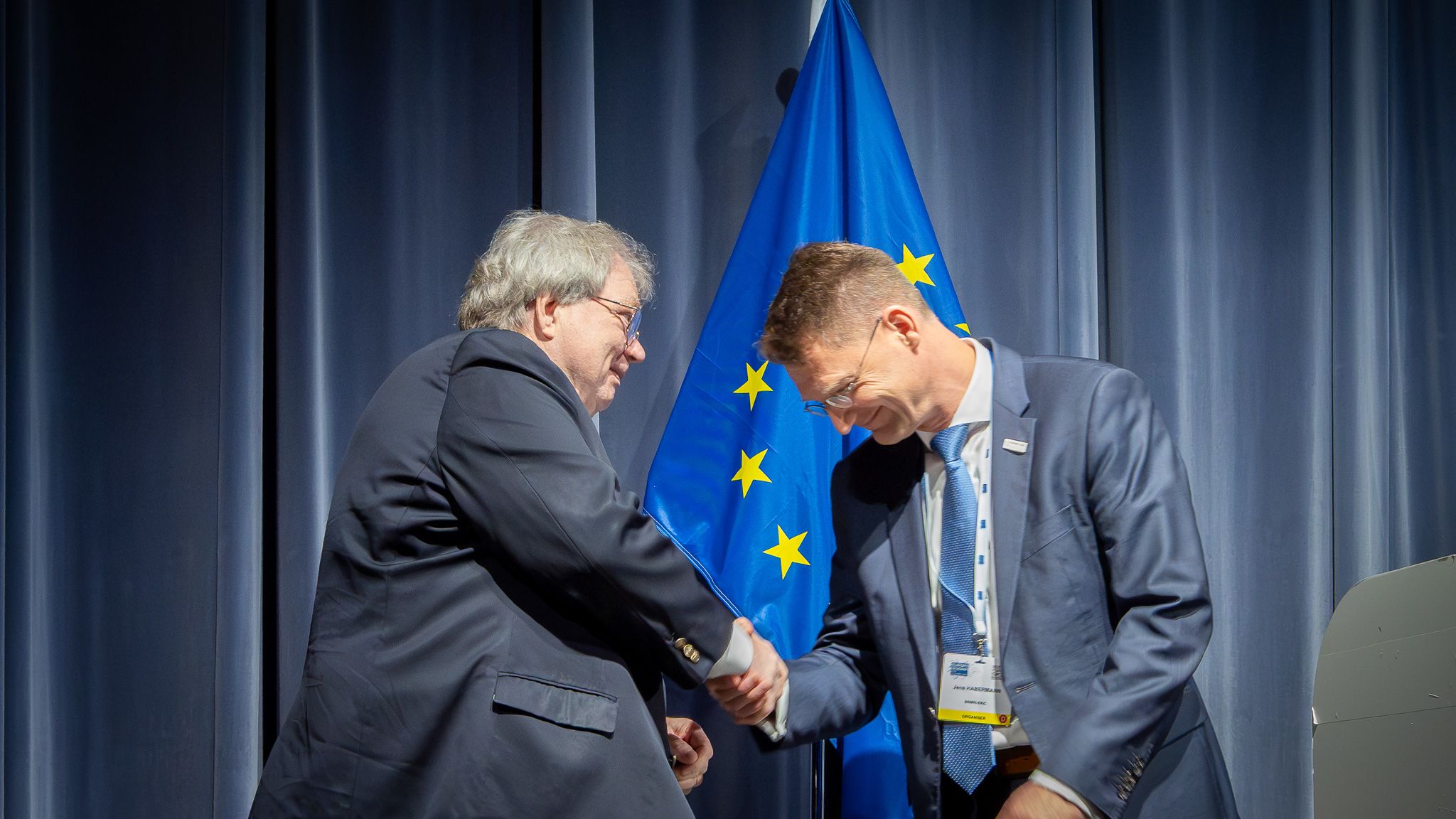 Two men shaking hands on stage in front of a curtain and an EU flag.