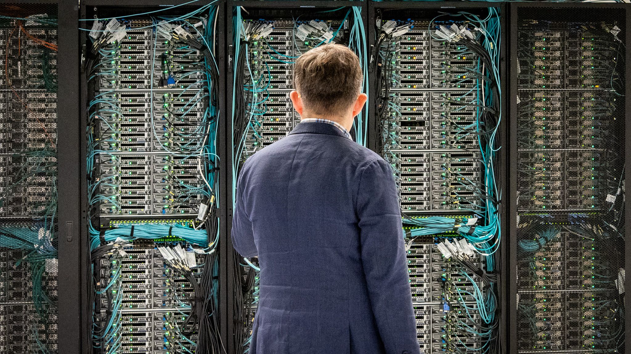 A man in a blue jacket looks at a large array of computer elements connected by cables inside a supercomputing centre.