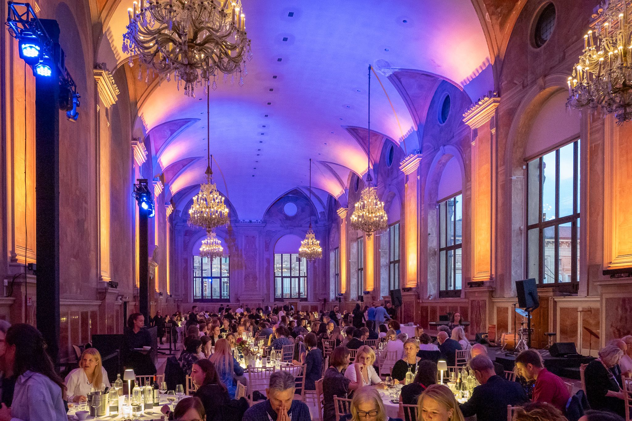 People in festive clothing sitting at large round tables in a colourful lit dining hall with chandeliers.