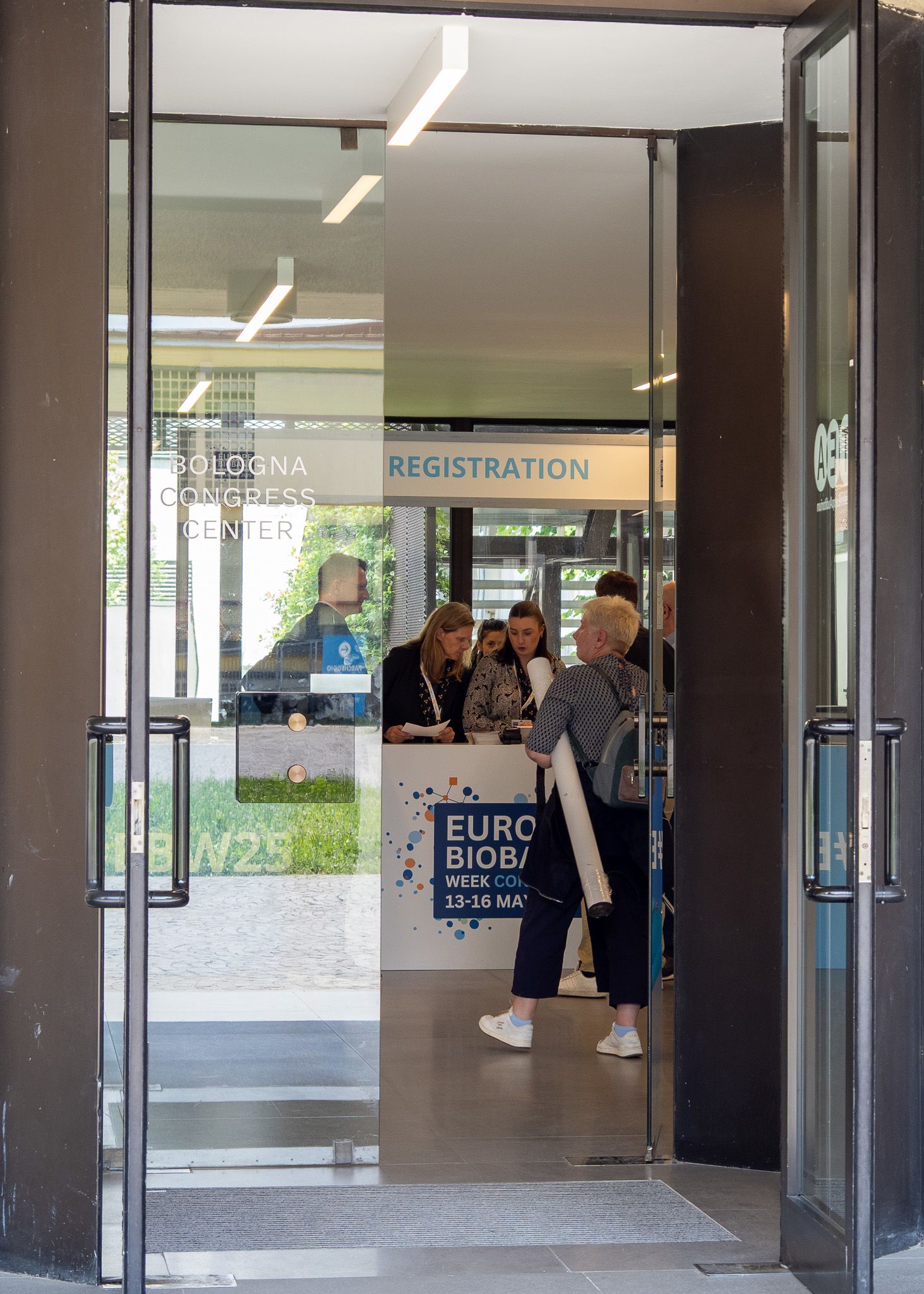 The image shows a group of people through a half open glass door standing at the registration desk of a congress centre.