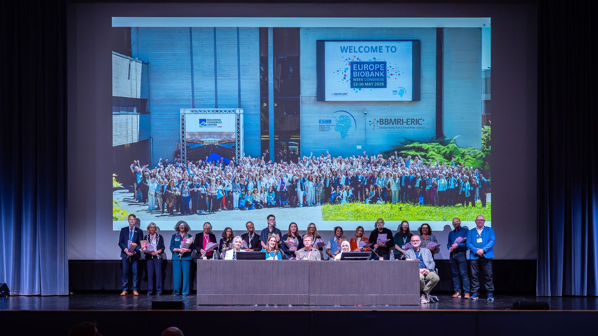 People with flowers on a stage in front of a large projection of a group photo of all EBW participants.